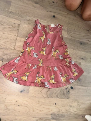 Rosa Bambi linnetopp från Disney Disney rosa klänning, storlek 68 - Supersöt rosa linnetopp från Disney med Bambi-tryck över hela plagget. Toppen har en volang nedtill och är ärmlös med rund hals. Materialet är mjuk bomull, perfekt för sommardagar. Passar dig som gillar gulliga och lekfulla prints!Söt klänning med Bambi-tryck. Klänningen har ett lekfullt mönster med Bambi och hans vänner.