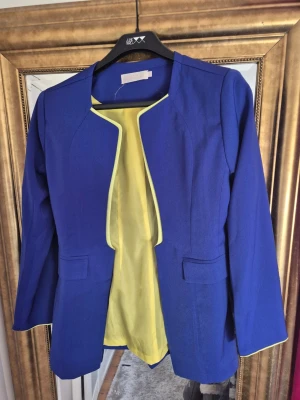 Blå kavaj med gul insida från Occzi - Snygg blå kavaj från Occzi med gul insida och gul kant längs öppningen. Kavajen har långärmad design, två fickor framtill och en stilren, öppen front utan knappar. Perfekt för dig som vill sticka ut med färg och stil.