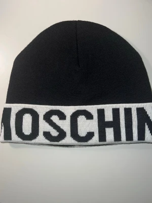 Moschino mössa - Säljer en stilren svart mössa från Moschino med bred vit kant och stor svart logotyp. Mössan är stickad och har en klassisk form som passar perfekt för att lyfta din look. Märket syns tydligt både på utsidan och insidan. Grym accessoar för dig som gillar designer. Mössan är typ aldrig använt 10/10 i skick!👊