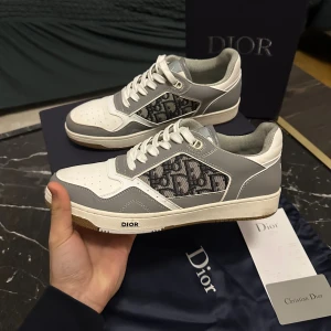 Dior sneakers i grått och vitt - Säljer ett par snygga Dior sneakers i grått och vitt med klassiskt Dior-mönster på sidan. Skorna har snörning, rund tå och platt sula. Materialet är en mix av skinn och textil, med detaljer i grått och vitt samt en beige yttersula. Perfekt för dig som gillar exklusiva märken och stilren design.