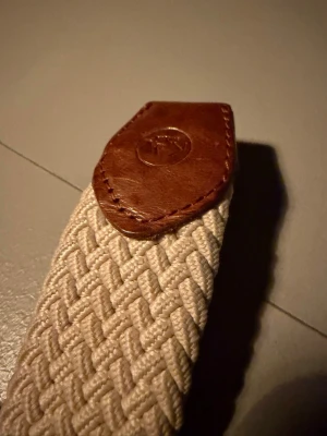 Beige flätad bälte med läderdetaljer - Snyggt beige flätat bälte med bruna läderdetaljer och metallspänne. Perfekt accessoar för att lyfta din outfit med en stilren och klassisk touch. Bältet har en robust känsla och passar till både jeans och chinos. Det är 110cm lång och bara att skriva om du har några funderingar 💬 Fraktar samma dag!