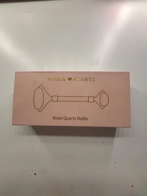 Rosenkvarts ansiktsroller - Säljer en lyxig ansiktsroller i äkta rosenkvarts med guldfärgade detaljer. Rollern har två stenar, en större och en mindre, perfekt för både ansikte och under ögonen. Handtaget är ljusrosa med guldfärgad text. Materialet är naturlig rosenkvarts och metall. Med en beskrivning om hur man kan rolla den i ansiktet.