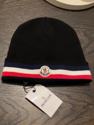 Svart Moncler mössa med ränder - Snygg svart mössa från Moncler med klassiska ränder i blått, vitt och rött längs kanten. Mössan har Moncler-logga framtill och är stickad i mjukt material. Perfekt accessoar för att lyfta din vinterstil.