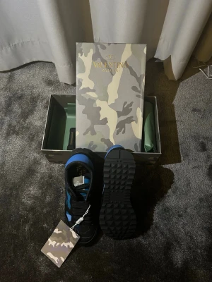 Valentino Garavani Rockrunner sneakers - Säljer ett par Valentino Garavani Rockrunner sneakers i svart och blått med coolt kamouflagemönster. Skorna har snörning, platt sula och rund tå. Materialet är en mix av skinn och textil. Kommer med originalkartong i matchande camo-design.