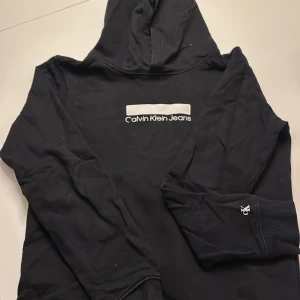 Svart hoodie från Calvin Klein Jeans - Svart hoodie från Calvin Klein Jeans med vit logotyp på bröstet och CK-detalj på ärmen. Tröjan har huva och ribbade muddar vid ärmslut och nederkant. Perfekt för dig som gillar stilrena och klassiska streetwear-plagg.