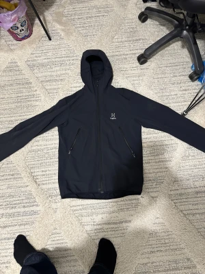 Svart vindjacka från Haglöfs med huva - Svart vindjacka från Haglöfs med stilren design, hel dragkedja framtill och två snedställda fickor med dragkedja. Jackan har en skön huva och är tillverkad i lätt syntetmaterial, perfekt för blåsiga dagar. Passformen är normal och ärmarna är långa.