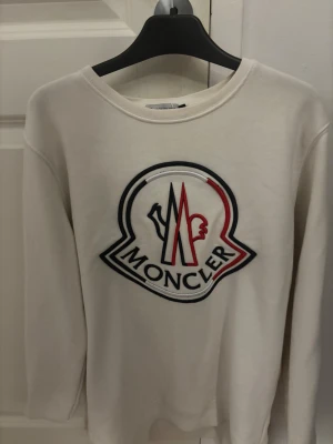 Vit sweatshirt från Moncler med stor logga - Vit långärmad sweatshirt från Moncler i bomull med stor broderad Moncler-logga i svart, rött och blått på bröstet samt mindre logga på ärmen. Rund hals och ribbade muddar. Perfekt för dig som gillar exklusiva streetwear-vibes.