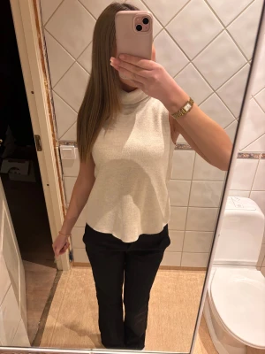 Beige polotopp i stickad material - Säljer en beige polotopp i mjukt stickat material. Toppen är ärmlös med hög krage och har en lätt utsvängd nederkant som ger en chill siluett. Perfekt att styla med svarta jeans eller kostymbyxor för en clean look. Pris kan doskuteras vid snabb affär
