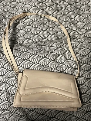 Beige axelväska i skinn - Snygg beige axelväska med stilren design och justerbart axelband. Väskan har ett lock med markerade sömmar och är tillverkad i slätt skinn. Perfekt för dig som vill ha en enkel men trendig accessoar till din outfit.