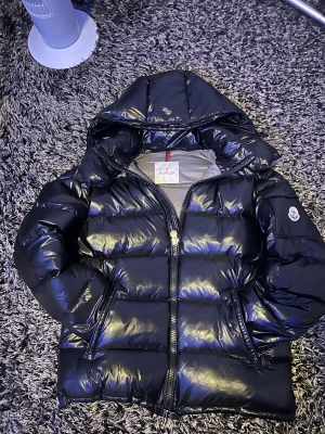 Svart Moncler pufferjacka med huva - Säljer en svart pufferjacka från Moncler med glansig finish och huva. Jackan har dragkedja framtill, två sidofickor med dragkedja och Moncler-logga på ärmen som scannar. Insidan har ett coolt serietryck och klassisk Moncler-etikett. Perfekt för kalla dagar.