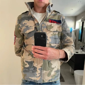 Zadig & Voltaire Camo Jacka - Snygg vindjacka i klassiskt camouflage från Zadig & Voltaire. Jackan har flera fickor framtill, patch på ärmen och coolt broderad detalj på bröstet. Dragkedja och knappar framtill, samt hög krage. Perfekt för dig som gillar streetstyle.
