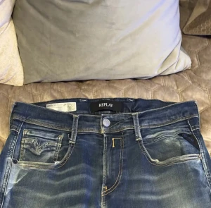 Replay Anbass jeans blå denim  - Snygga Replay Anbass jeans i klassisk blå tvätt med slitningar och lätt distressed detalj på ena benet. Modellen har fem fickor, kontrastsömmar och normal passform. Nypris 1499 mitt pris 499
