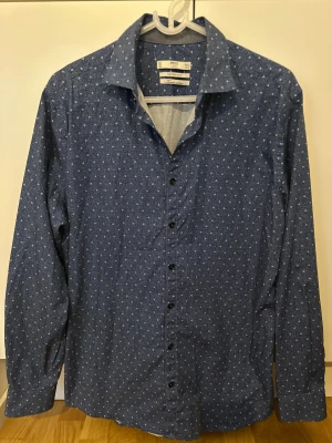 Mango - Blå mönstrad skjorta från MNG Man - Snygg blå skjorta från Mango Man i modern slim fit med små vita prickar i ett diskret mönster. Skjortan har långa ärmar, klassisk krage och svarta knappar framtill. Tillverkad i ett mjukt bomullstyg som känns skönt mot huden. Perfekt för dig som gillar stilrena detaljer.