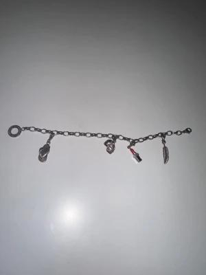 Silverfärgat Thomas Sabo charmarmband med berlocker - Charmigt armband i silverfärgad metall med fyra berlocker: flipflop, dödskalle, läppstift i rött och svart samt en fjäder. Kedjan har stora länkar och ett klassiskt lås. Perfekt accessoar för dig som gillar unika detaljer och personlig stil.
