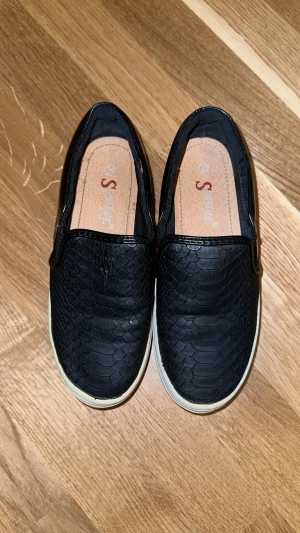 Svarta slip-on sneakers med reptilmönster 36  - Säljer ett par svarta slip-on sneakers med coolt reptilmönster på ovandelen. Skorna har en vit platt sula och är tillverkade i syntetmaterial med en blank finish runt kanten. Perfekt för dig som gillar stilrena och trendiga skor.