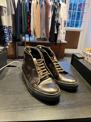 Acne Studios Mörkbruna höga sneakers i skinn - Snygga mörkbruna höga sneakers med rund tå och platt sula. Skorna är tillverkade i skinn och har matchande bruna snören. På sidan finns en präglad logga. Insidan är vadderad för extra komfort och sulan är svart med vågigt mönster undertill.