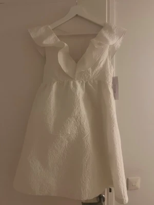 Vit kortklänning med volang från Bubbleroom - Supersöt vit kortklänning från Bubbleroom med v-ringning och bred volangkant upptill. Klänningen har ett strukturerat tyg med blommig brodyr och är ärmlös. Perfekt för sommarens alla tillfällen när du vill känna dig extra fin. Är helt ny med prislapp på och i storlek xs