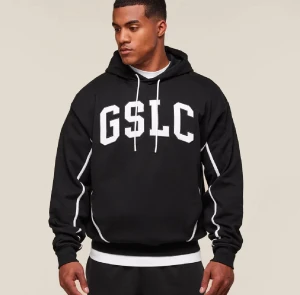 Svart hoodie gymshark  - Svart hoodie med stor vit GSLC-text på bröstet och vita detaljer längs ärmarna. Tröjan har huva med snörning och ribbade muddar vid ärmslut och nederkant. Perfekt för dig som gillar sportig och avslappnad stil.