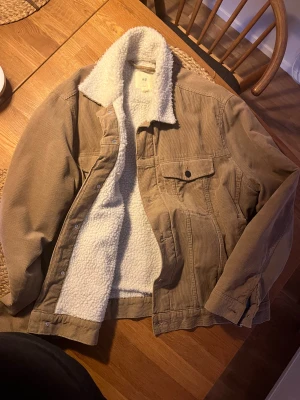 Beige teddyjacka från H&M - Säljer en beige teddyjacka  från H&M med mjukt vitt foder och krage i teddy. Jackan är gjord i manchestertyg och har klassiska knappar framtill samt fickor med lock. Perfekt för dig som gillar chill och snygg stil under höst och vinter.