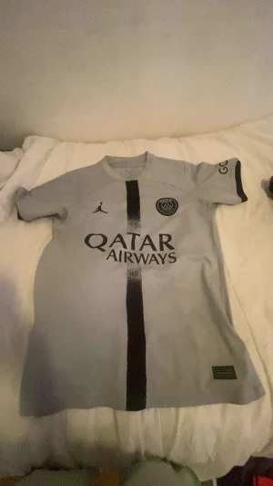 PSG x Jordan grå fotbollströja - Snygg grå PSG x Jordan t-shirt med svart vertikal rand och tryck 'Qatar Airways' på bröstet. Kortärmad modell med rund hals och PSG-logga på vänster sida. Perfekt för dig som älskar fotboll och vill ha en unik tröja med stilren design. Neymar på baksidan