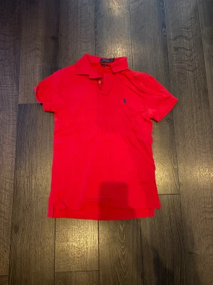 Röd piké från Polo Ralph Lauren - Säljer en röd pikétröja från Polo Ralph Lauren i slim fit-modell. Klassisk krage, korta ärmar och blå broderad logga på bröstet. Materialet är mjuk bomull som känns skönt mot huden. Perfekt för dig som gillar stilren och sportig look.