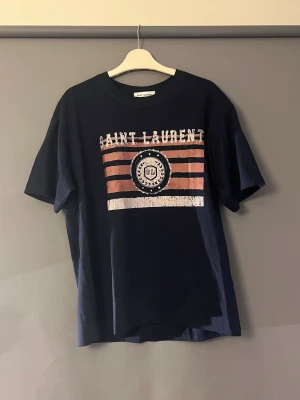 Mörkblå Saint Laurent t-shirt - Snygg mörkblå t-shirt från Saint Laurent med tryck i vitt och rosa på bröstet. T-shirten har korta ärmar och klassisk rund hals. Materialet är mjuk bomull och passformen är normal. Helt ny, beställde fel storlek.