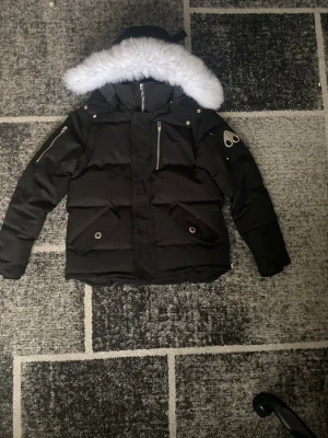 Svart Moose Knuckles dunjacka med päls - Säljer en svart dunjacka från Moose Knuckles med vit päls på huvan. Jackan har flera dragkedjor, knappar och logotyp på ärmen. Fodrad insida med broderad patch och innerficka. Perfekt för kalla dagar och har en riktigt snygg streetstil.