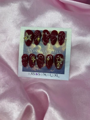 Ryx (S);Short Almond;Curved - Snygga handgjorda vinröda press-on naglar med glittereffekt och coola guldstjärnor, pärlor och 3D handmålad linjeart. Naglarna har en kort almond form och kommer i S: 15 mm, 11,5 mm, 12 mm, 11 mm, 9 mm. Perfekt för dig som vill ha unika och trendiga naglar utan att gå till salong.