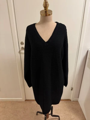 Svart stickad klänning Missguided - Mysig svart stickad klänning från Missguided med v-ringning och lång ärm. Oversized passform och ribbad struktur, perfekt för en chill och trendig look. Modellen är längre och passar dig som är lång.