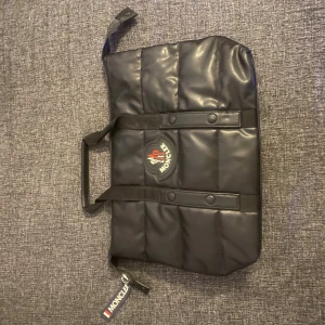 Svart quiltad datorväska från Moncler - Snygg svart quiltad datorväska från Moncler med klassisk logga framtill och coolt serietidningsfoder på insidan. Väskan har dragkedja, dubbla handtag och Moncler-tag på sidan. Perfekt för dig som vill ha en stilren och trendig accessoar.