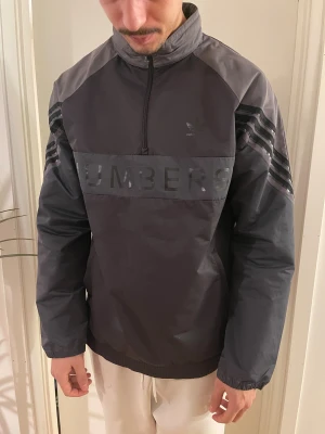 Svart vindjacka Adidas Skateboarding - Säljer en svart och mörkgrå vindjacka från Adidas Skateboarding 'NUMBERS' på bröstet och klassiska tre ränder på ärmarna. Jackan har half zip, sidofickor med dragkedja och elastisk nederkant. 