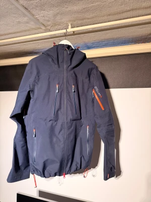 Stellar equipment shell jacket - Mörkblå skaljacka från Stellar Equipment med alla funktioner man kan behöva för skidåkning eller andra friluftsaktiviteter.  Jag har huvudsakligen använt den för utförsåkning. Det finns vissa tecken på användning, det mest tydliga är på framsidan där tyget är lite smält från att jag ramla på en rail. Som man ser på bilderna är skadan bara ytligt vilket gör att skalet fortfarande fungerar. Visar bara på att åkaren är seriös😅 Annars fint skick, endast använd en vinter.  Jackan är fullt utrustad fö