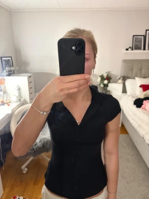 Svart figurnära blus med knappar - Säljer en svart, figurnära blus med korta ärmar och knappar hela vägen framtill. Blusen har en liten ståkrage och är sydd i ett mjukt, stretchigt material som formar sig snyggt efter kroppen. Från Shein men skönt material. Aldrig använt. Storlek S 