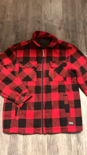 Rutig röd overshirt från Bluewear - Säljer en röd och svart rutig overshirt från Bluewear i storlek Large. Jackan har två bröstfickor med knappar, dragkedja framtill med läderdetalj och quiltat svart foder. Perfekt för lager-på-lager och chill stil.