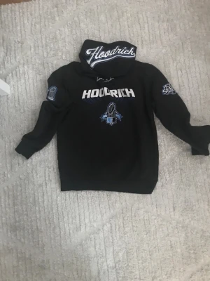 Svart Hoodie från Hoodrich med tryck - Svart hoodie från Hoodrich med stora vita och blå tryck både fram och bak. Hoodien har huva med broderad logga, ribbade muddar och flera coola patchar på ärmarna. Perfekt för dig som gillar streetwear och vill sticka ut med snygga detaljer.