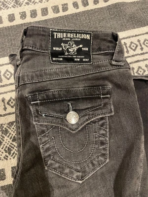 True Religon bootcut jeans - Ett par snygga grå-svartajeans med detaljer på fickorna och en bootcut modell. Omkretsen vid midjan: 68cm. Innerbenets längd: 84cm.