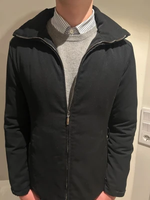 Svart Moncler dunjacka med dragkedja - Svart dunjacka från Moncler med quiltad insida och stilren design. Jackan har hög krage, hel dragkedja framtill och långärmade ärmar. Materialet är polyester och insidan är dunfylld för extra värme. Perfekt för dig som vill ha en snygg och funktionell jacka.