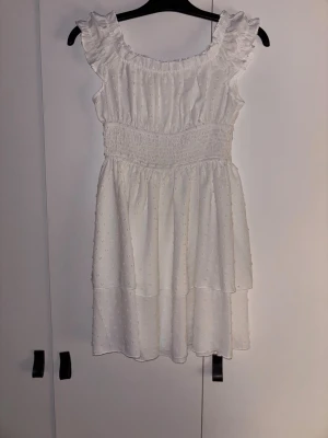 Vit kort sommarklänning med volang - Supersöt vit kortklänning med volangdetaljer vid axlarna och smockad midja. Klänningen har ett diskret prickigt mönster och är ärmlös med en luftig, lätt känsla. Perfekt för varma dagar och har en romantisk vibe.