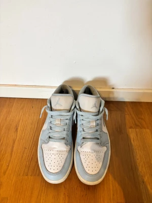 Nike Air Jordan 1 Low ljusblå/vit - Nike Air Jordan 1 Low sneakers i ljusblått och vitt med klassisk snörning och rund tå. Skorna har en platt sula och är tillverkade i skinn och syntet. Swoosh-logga på sidan och Jumpman på tungan ger en ikonisk look. Perfekt för dig som gillar streetwear och sneakers.