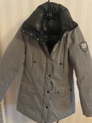Grå och svart vändbar pufferjacka st S - Säljer en vändbar pufferjacka i grått och svart. Ena sidan är grå med diskret randigt mönster och snöflinge-patch på ärmen, den andra sidan är helsvart och glansig. Jackan har tryckknappar, dragkedja och stora fickor. Perfekt för dig som vill ha två stilar i en jacka. Kan endast knäppas med knappar, dragkedja funkar dock saknas hak som man drar i , kan kosta mellan 50-80 kr hos syaffärer eller tas från nån annan jacka.