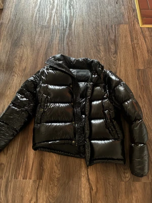 Svart pufferjacka från Peak Performance - Säljer en svart, glansig pufferjacka från Peak Performance. Jackan har hög krage, dragkedja och tryckknappar framtill samt två fickor med knappstängning. Perfekt för kalla dagar och riktigt snygg till streetwear-stilen.