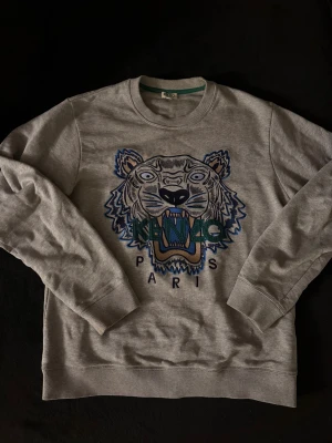 Grå Kenzo Tröja - Grå stilren tröja från Kenzo med ikonisk broderad tiger på bröstet och texten 'KENZO PARIS' i grönt och blått. Tröjan har rund hals, ribbade muddar och är tillverkad i mjuk bomull. Perfekt för dig som gillar statement-plagg och streetstyle.