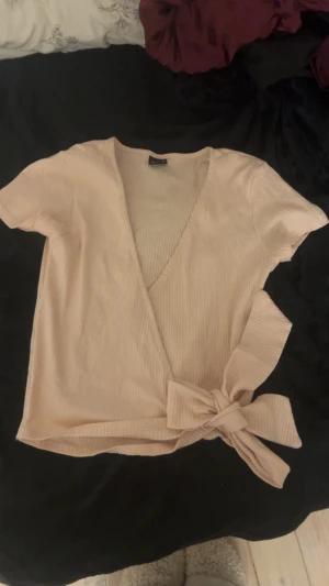 Ljusrosa omlottopp med rosett - Säljer en ljusrosa omlottopp med korta ärmar och snygg v-ringning. Toppen har ribbad struktur och en stor rosett på sidan som ger extra stilpoäng. Perfekt för dig som gillar en chill men ändå trendig look.