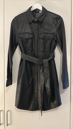 Svart skinnklänning från Amisu - Säljer en svart kort klänning i skinnimitation från Amisu. Klänningen har långa ärmar, knappar framtill, två bröstfickor och ett matchande bälte i midjan. Snygg skjortkrage och coola detaljer som ger en edgy look.