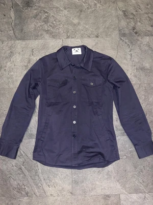 Mörkblå overshirt från Stilgruvan - Snygg mörkblå overshirt från Stilgruvan i 100% bomull. Klassisk skjortkrage, två bröstfickor med knappar och långa ärmar med manschett. Perfekt lager-på-lager-plagg med stilrena knappar framtill. Nypris 500kr, knappt andvänd!