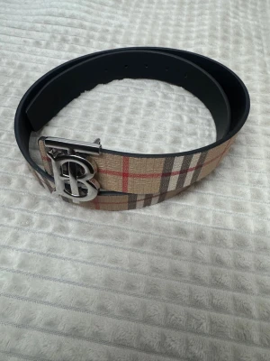 Rutig Burberry bälte med silverdetalj - Snyggt bälte från Burberry med klassiskt rutigt mönster i beige, rött, svart och vitt. Bältet har en bred design och en stor silvrig spänne med Burberrys ikoniska TB-logga. Tillverkat i skinn med tygdetaljer på ovansidan.