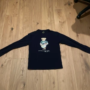 Mörkblå långärmad Polo Bear-tröja - Mörkblå långärmad tröja från Polo Ralph Lauren med ikoniskt Polo Bear-tryck på bröstet. Tröjan har en klassisk passform och är tillverkad i mjuk bomull, perfekt för dig som gillar stilrena och lekfulla detaljer.