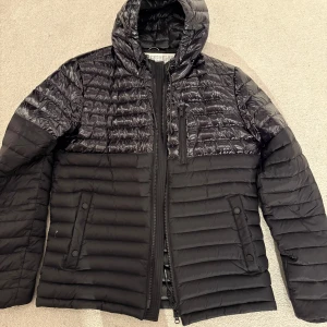 Svart pufferjacka från Moose Knuckles - Säljer en svart pufferjacka från Moose Knuckles med glansig finish upptill och matt nedtill. Jackan har huva, dragkedja, flera fickor och diskret logotyp på ärmen. Perfekt för kyliga dagar.