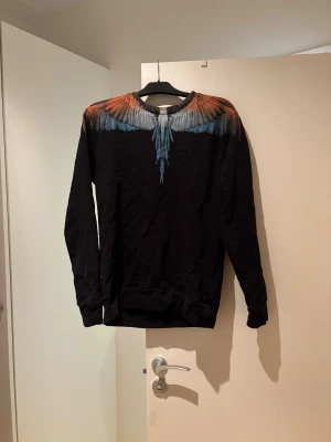 Svart tröja med vingar från Marcelo Burlon - Svart långärmad tröja från Marcelo Burlon County of Milan med ikoniskt vingtryck i orange, blått och grått över axlar och hals. Tröjan har rund hals och ribbade muddar. Perfekt för dig som vill sticka ut med en unik design.