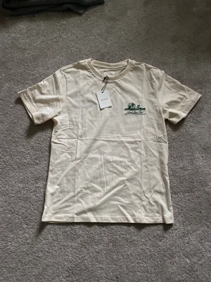 Aimé Leon Dore creme t-shirt med tryck - Snygg beige t-shirt från Aimé Leon Dore med grönt grafiskt tryck på bröstet och ryggen. Klassisk rund hals och korta ärmar. Mjuk bomullskvalitet som känns skön mot huden. Perfekt för dig som gillar streetwear och stilrena prints. Nyrpis 875kr